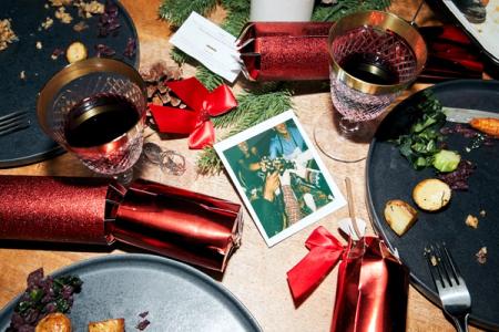 A festive table after a meal, with plates of food remnants, wine glasses, Christmas crackers, and a polaroid photo.