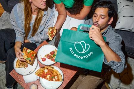 Three people sharing pizzas, one man looking into a green "Too Good To Go" bag.