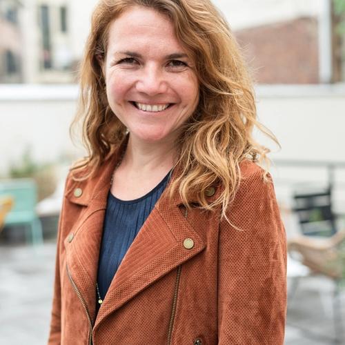 Smiling woman with wavy reddish-blonde hair wearing a brown jacket outdoors.