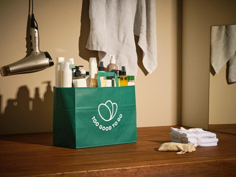 A green Too Good To Go bag filled with toiletries and beauty products on a wooden counter, with a hairdryer and towel in the background.