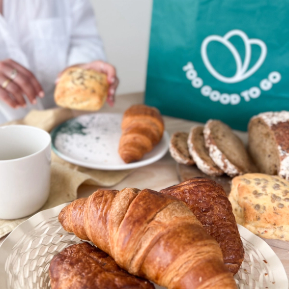 a croissant sits on a plate next to a bag that says too good to go