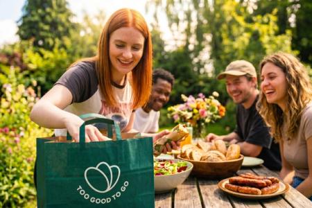 A woman unpacks food from a green "Too Good To Go" bag at an outdoor meal with smiling friends.