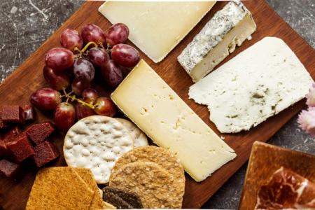A wooden board featuring various cheeses, red grapes, crackers, quince paste, and prosciutto.