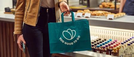 A person holds a green "Too Good To Go" branded bag in a cafe.