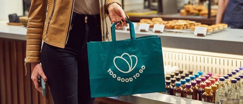 A person holds a green "Too Good To Go" branded bag in a cafe.