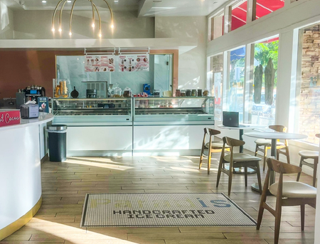 Interior of a bright ice cream shop with display cases, a white counter, and a seating area by large, sunny windows.