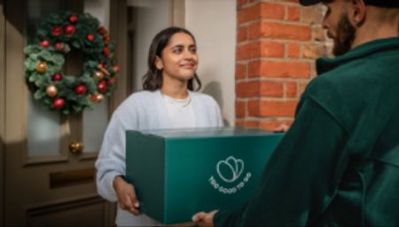 A smiling woman receives a green Too Good To Go box from a delivery person at a doorway with a Christmas wreath.