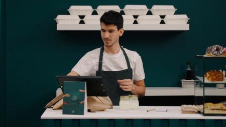 A man in a dark apron works at a cafe counter, preparing takeout orders with bags, jars, and a tablet, with takeout containers on a shelf above.