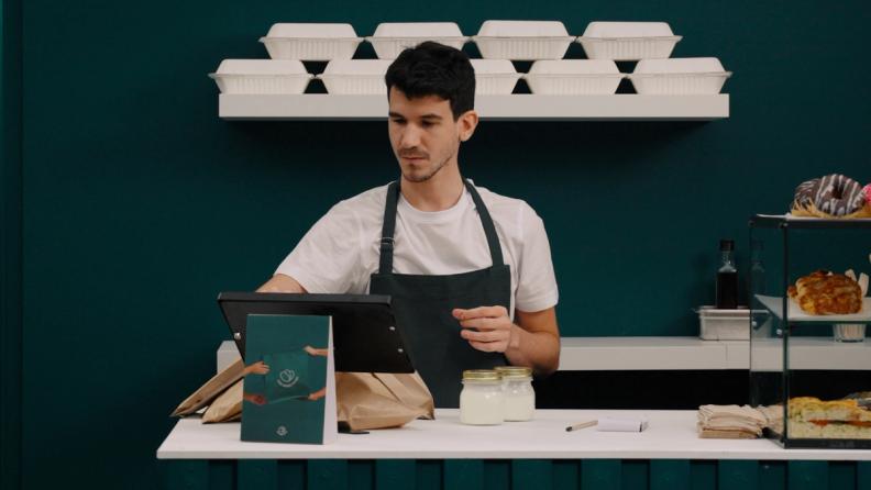 A man in a dark apron works at a cafe counter, preparing takeout orders with bags, jars, and a tablet, with takeout containers on a shelf above.