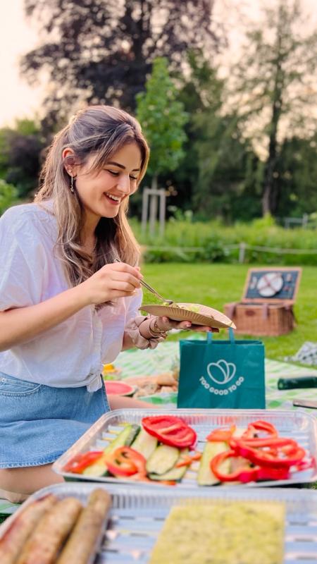 a woman sitting on a picnic blanket with a bag that says " too good to go "