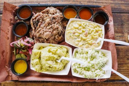 A BBQ platter with pulled pork, mac and cheese, potato salad, coleslaw, pickles, and various sauces.
