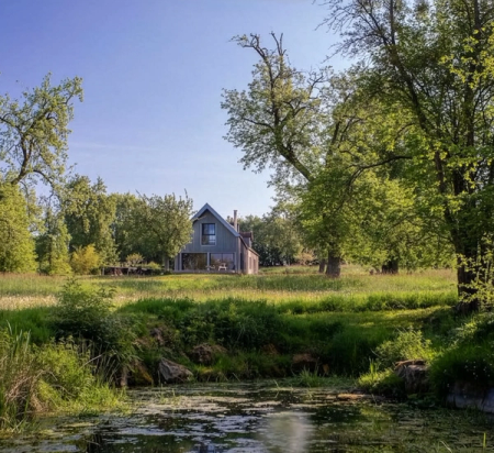 A modern gabled house with large windows stands in a green field, bordered by trees, with a pond in the foreground.