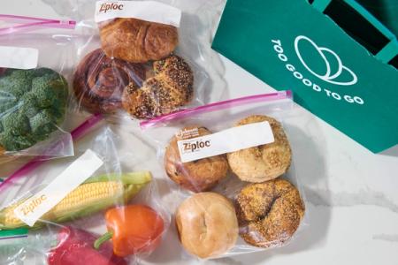 Broccoli, corn, bell peppers, pastries, and bagels packaged in Ziploc bags, next to a green "Too Good To Go" bag.