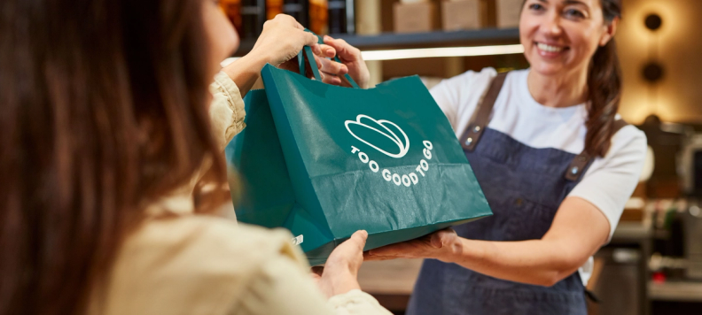 Woman wearing an apron handing over a Too Good To Go bag
