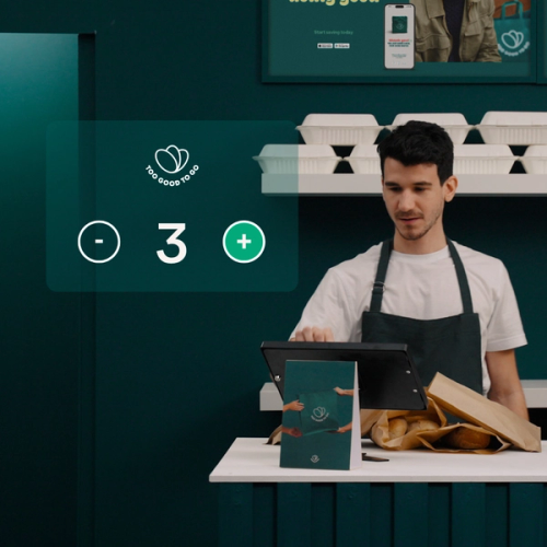 a man behind a counter with a sign that says Too Good To Go