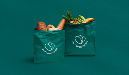 Two green "Too Good To Go" bags, one filled with bread and pastries, the other with fruits and vegetables, on a teal background.