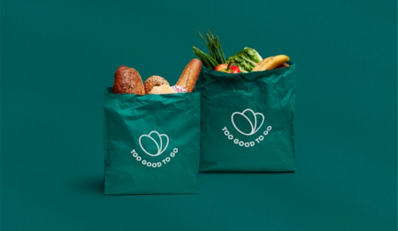 Two green "Too Good To Go" bags, one filled with bread and pastries, the other with fruits and vegetables, on a teal background.