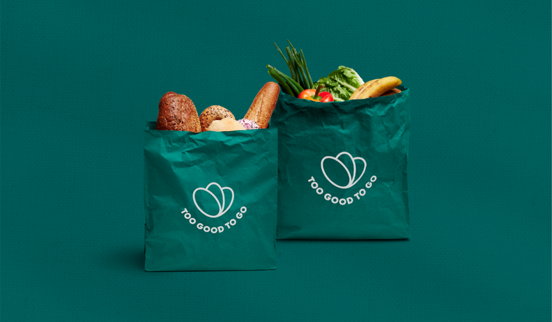 Two green "Too Good To Go" bags, one filled with bread and pastries, the other with fruits and vegetables, on a teal background.