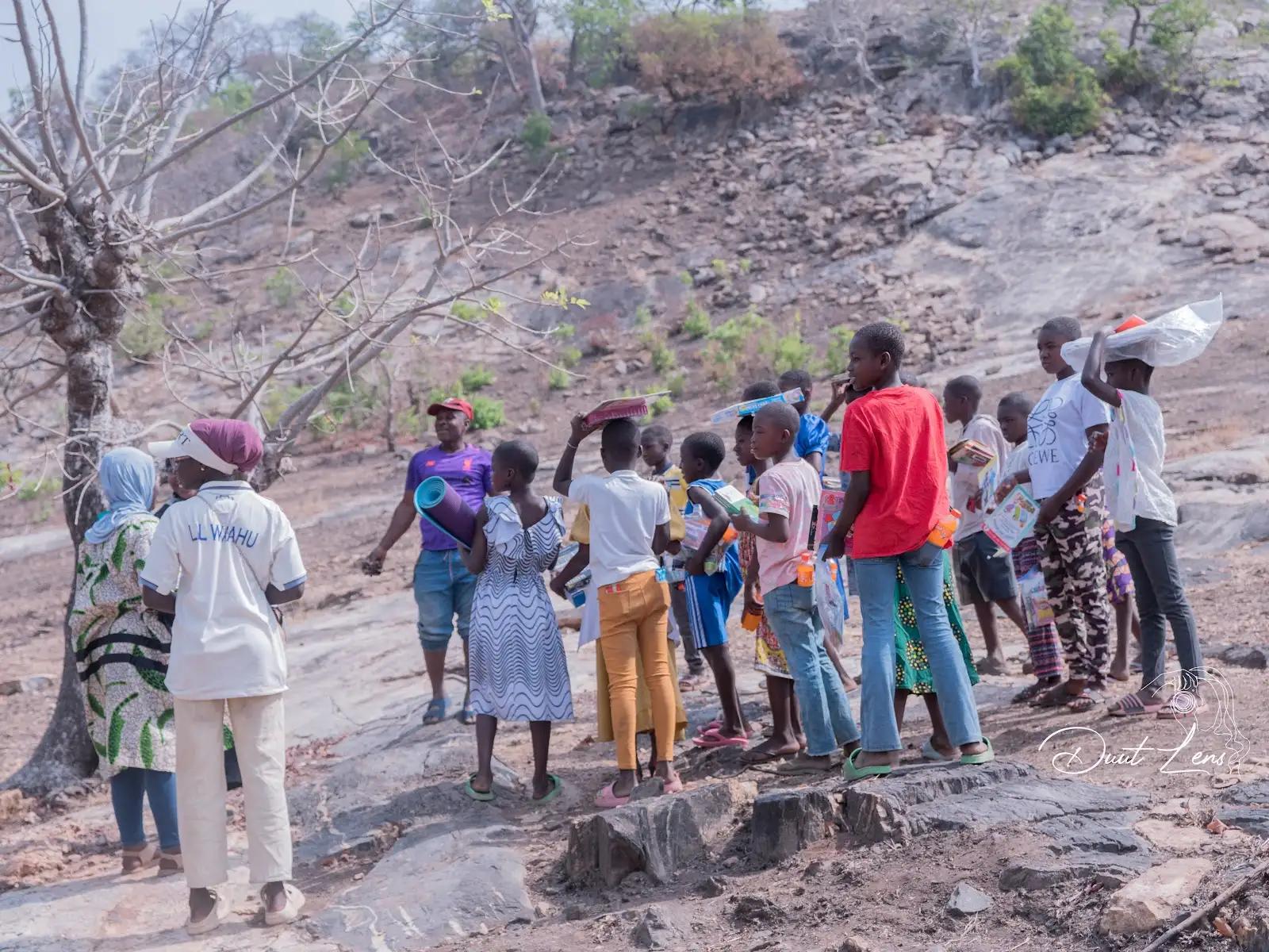 Children and adults walking on rocky Uombo Rock terrain, carrying books and bags