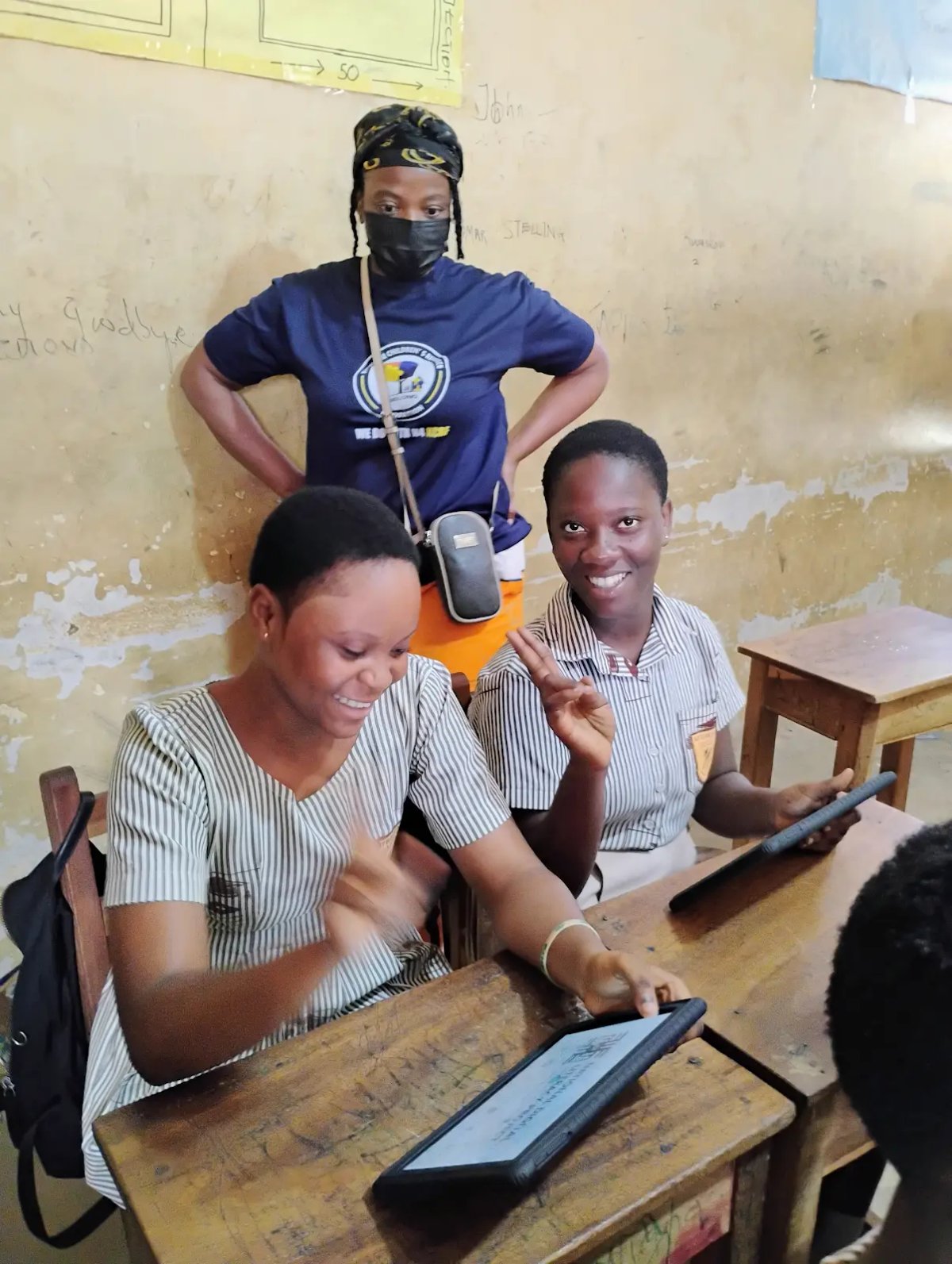 Two girls smiling while working on a tablet in their classroom, supervised by KAPAW staff