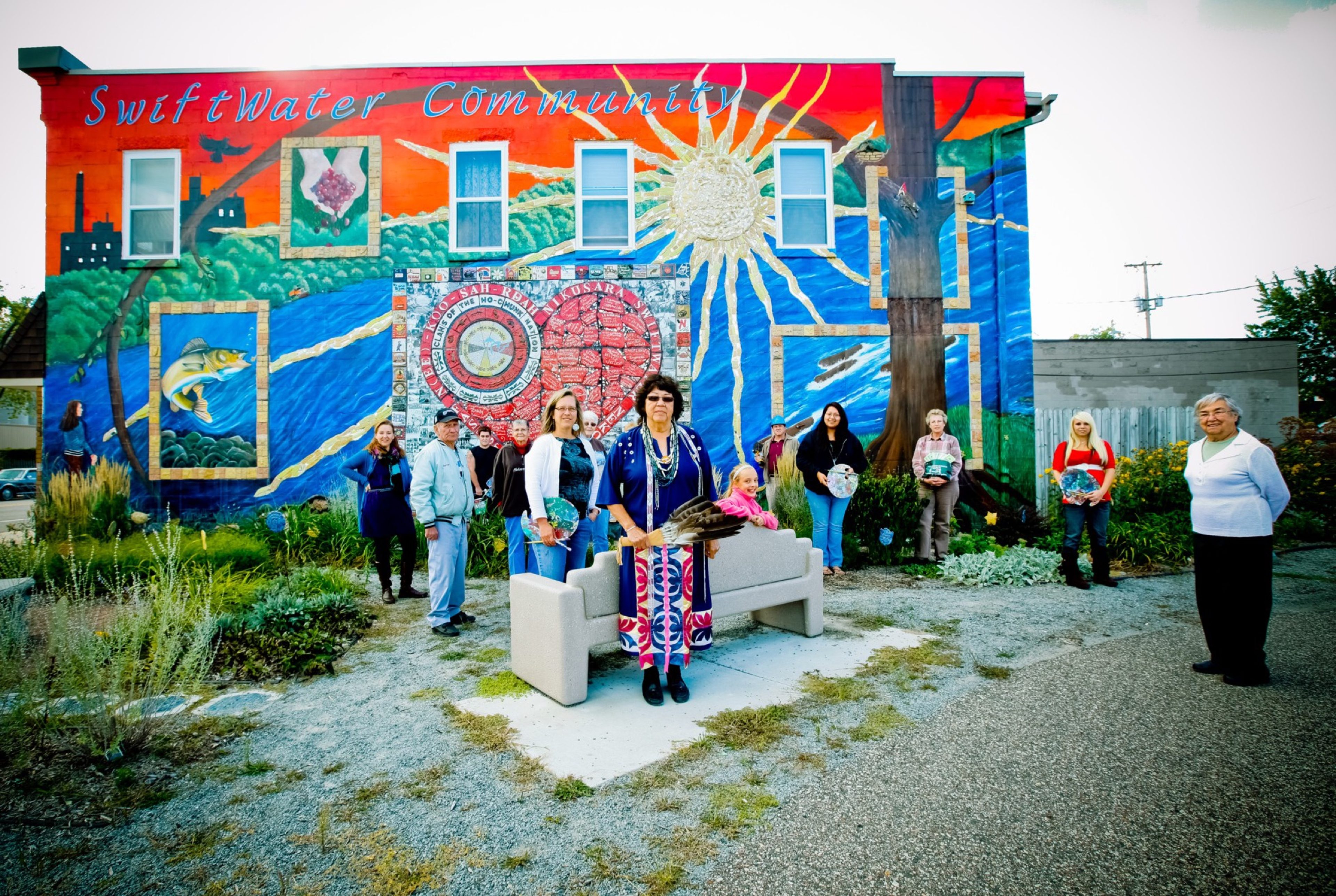 Community members gathered around a bench in front of the SwiftWater Community mural, a sunburst and river painted across a brick wall.