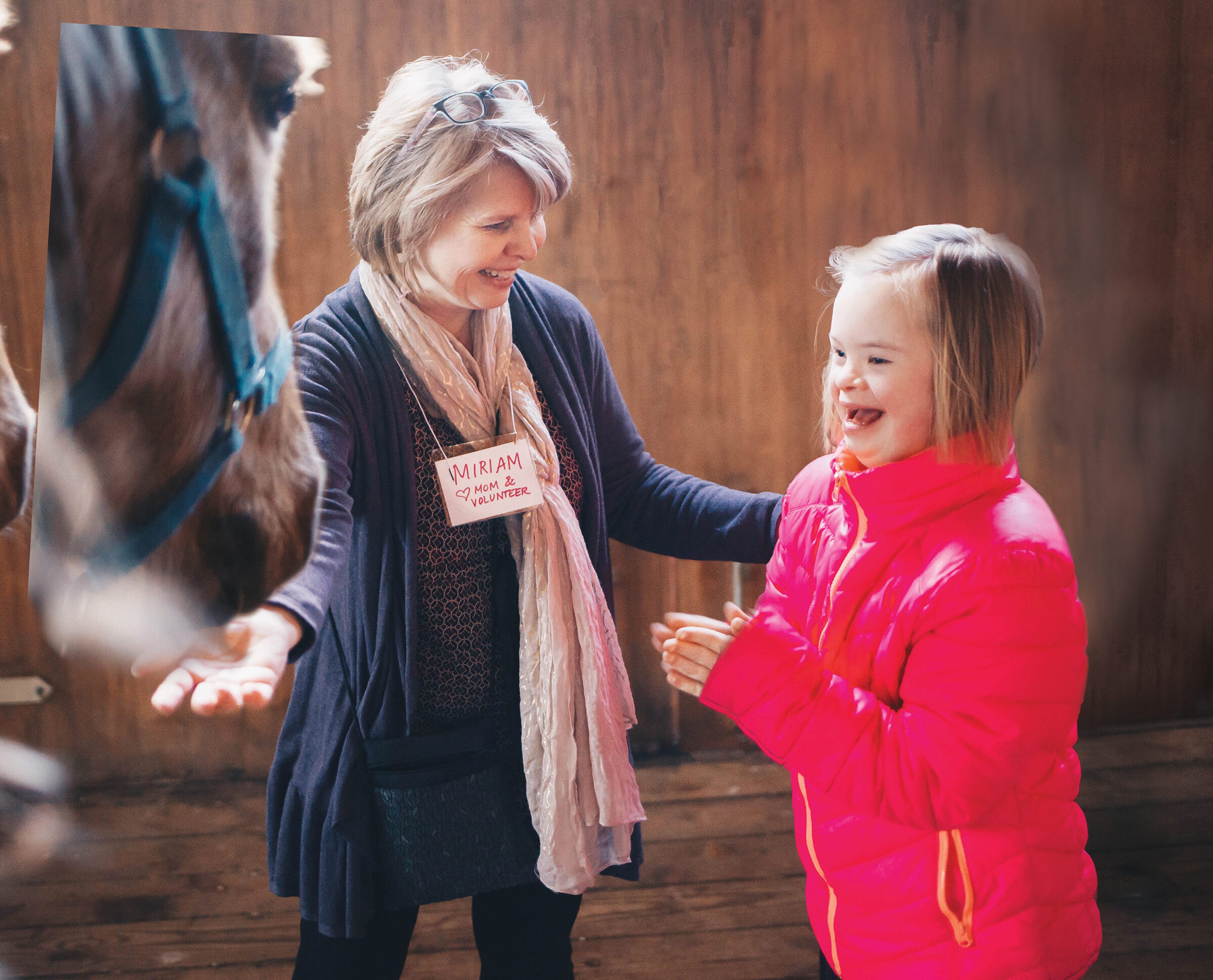 A volunteer in a name tag reading Miriam, mom and volunteer, laughing with a delighted girl beside a horse in a barn.