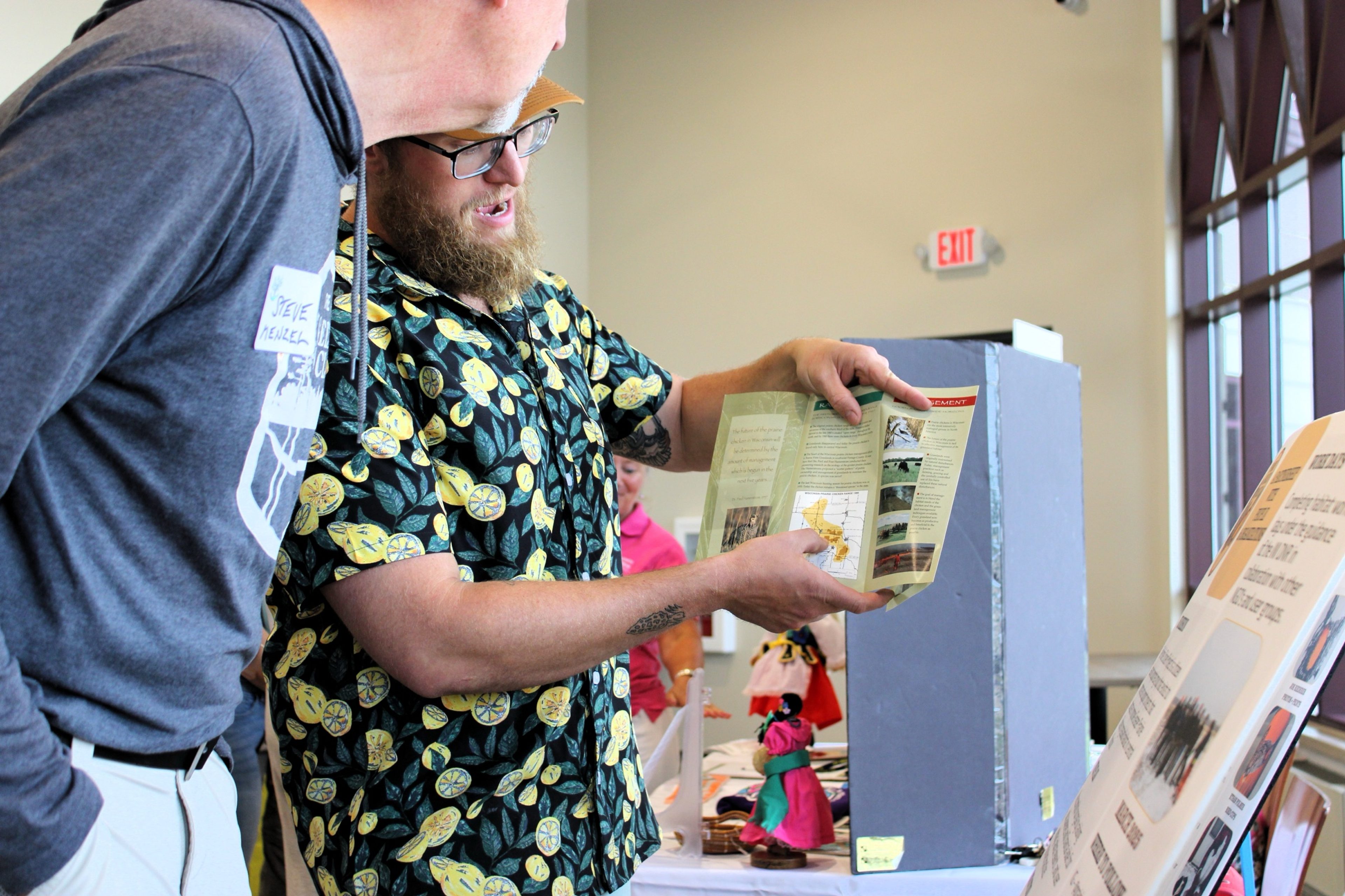 A man in a lemon-print shirt reading a brochure at a display table during a community event.