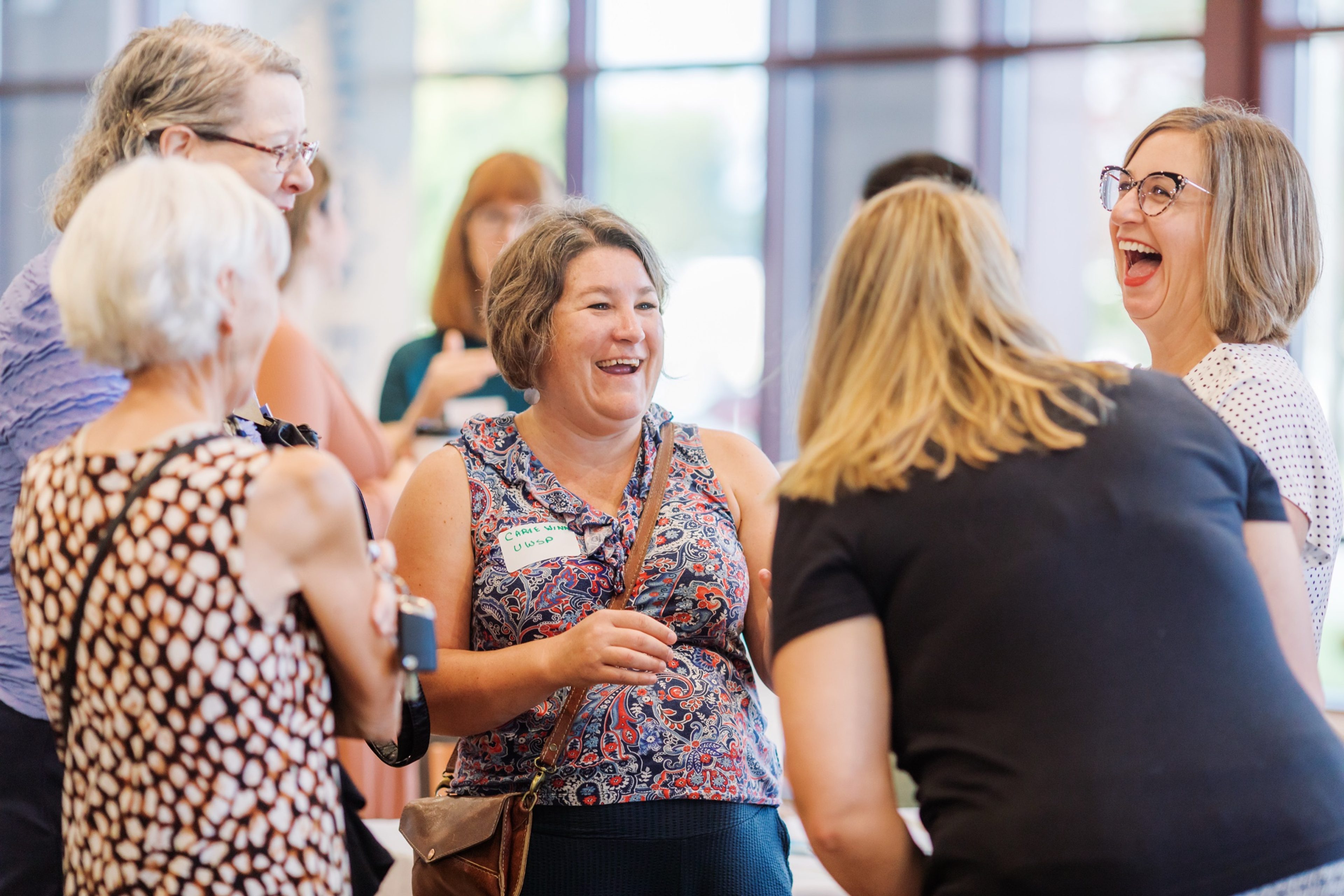 A group of women laughing together in a bright, glass-walled lobby at a community event.