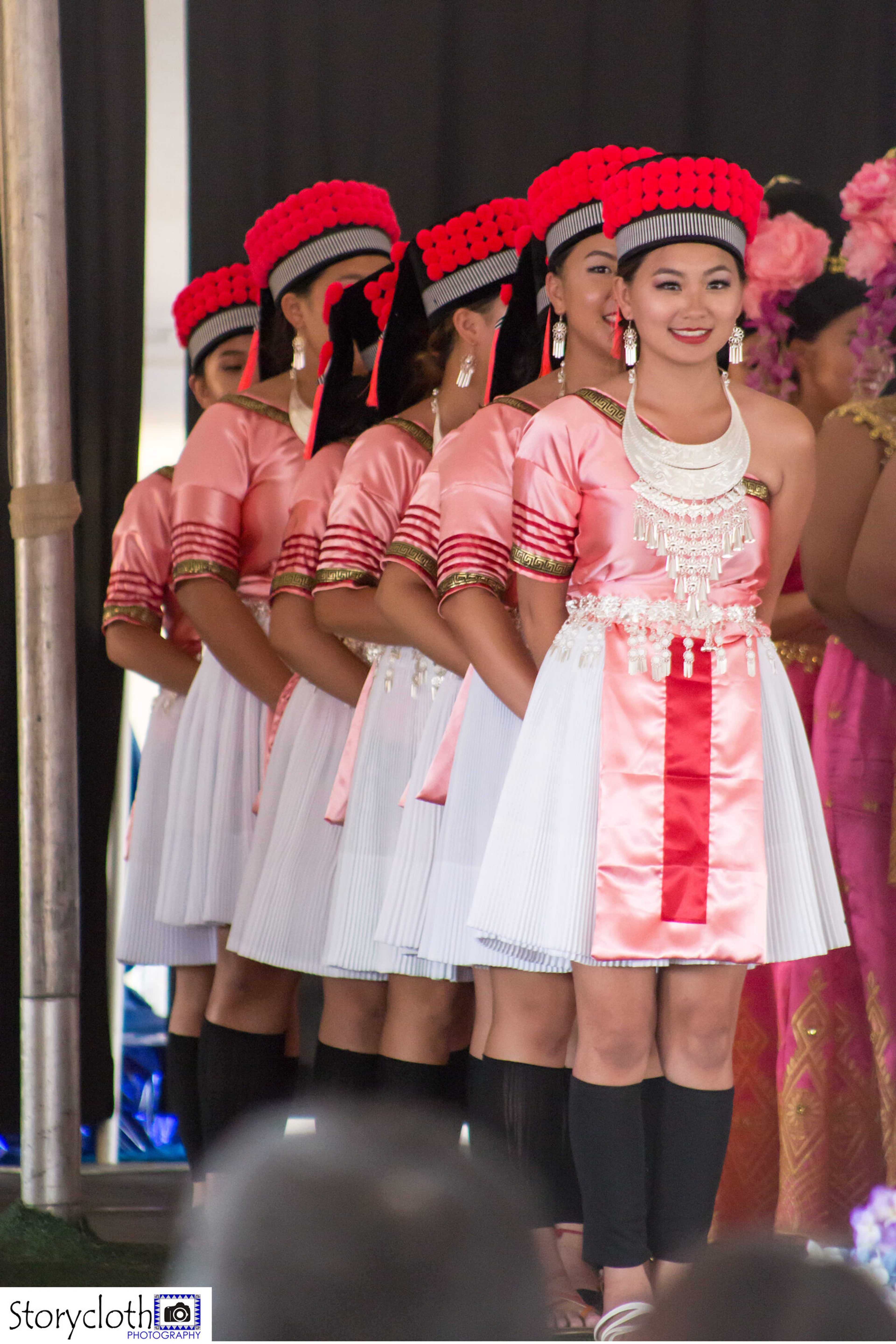Dancers in pink and white Hmong dress with red pom headpieces lined up on stage at a community festival.