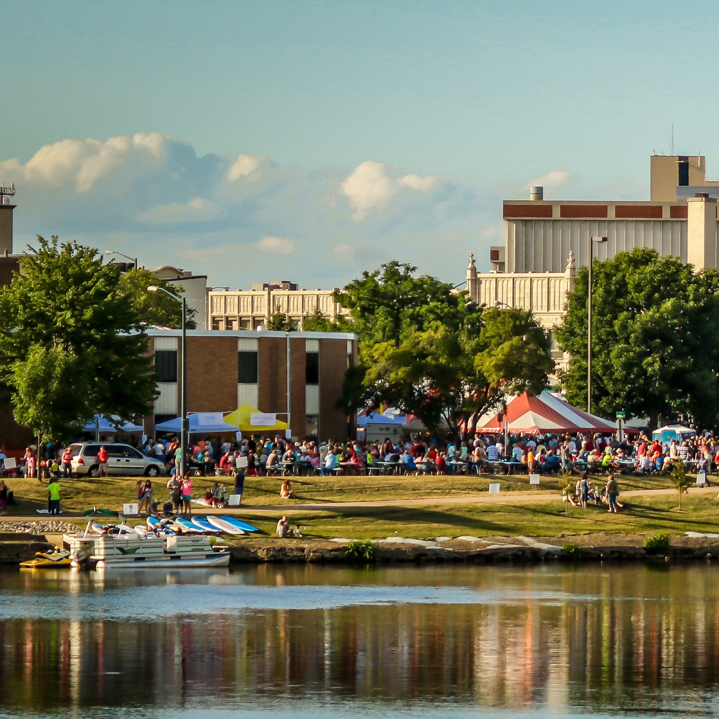 A riverfront festival crowd under striped tents on the lawn in Wisconsin Rapids, pontoon boats tied up along the bank.