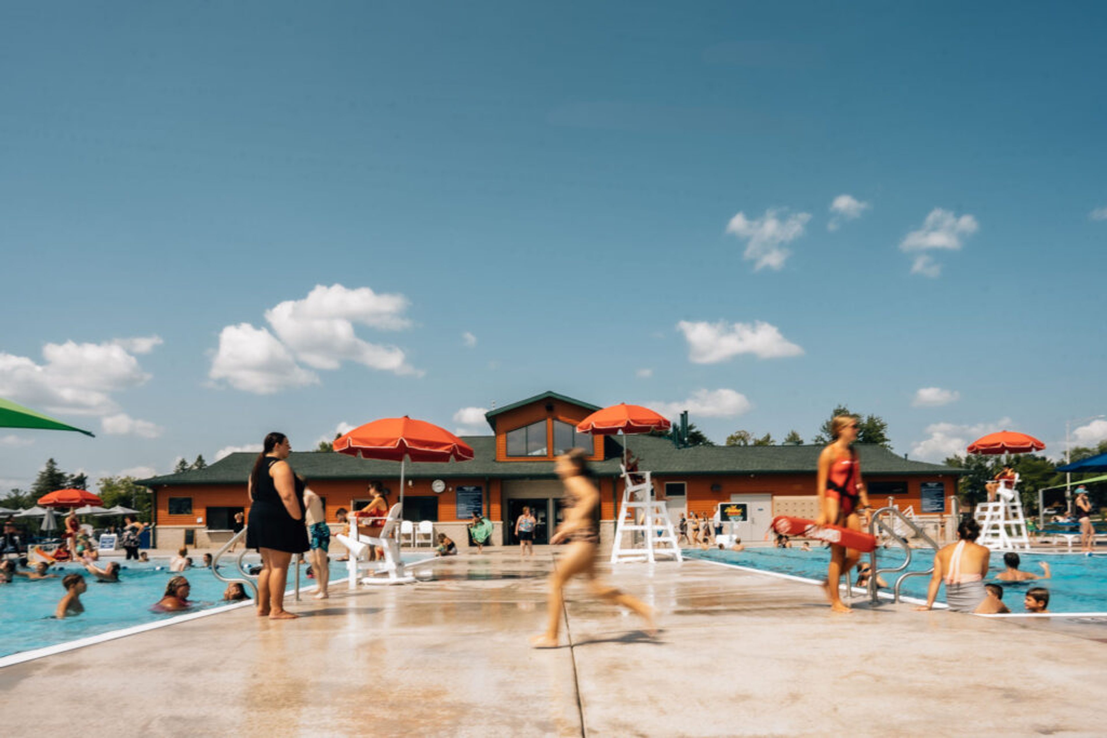A child running along the wet deck of a busy municipal pool, red umbrellas and lifeguard chairs lining the water in front of an orange bathhouse.