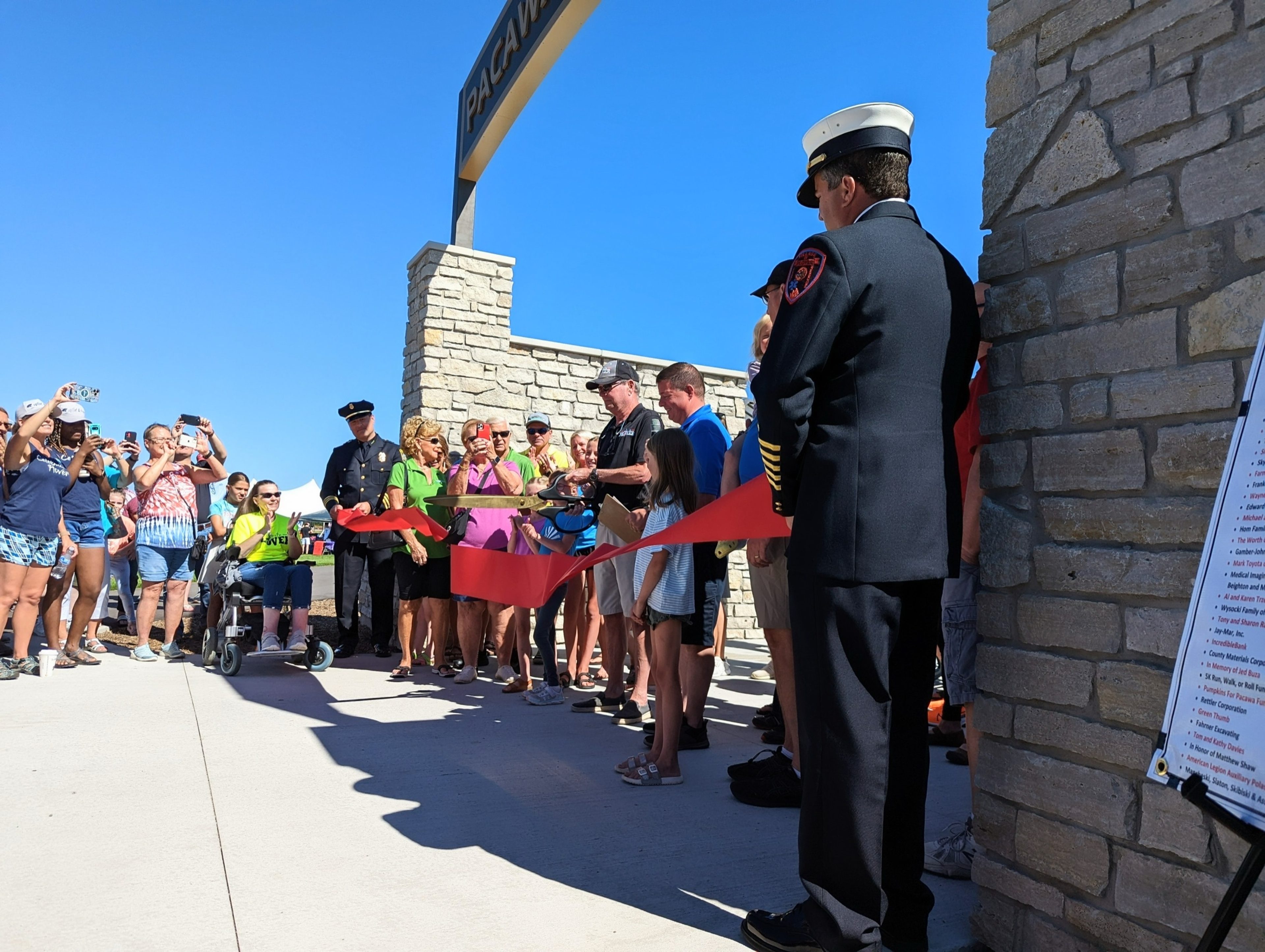 A fire chief in dress uniform watching neighbors cut a red ribbon at a new stone park entrance under a clear blue sky.