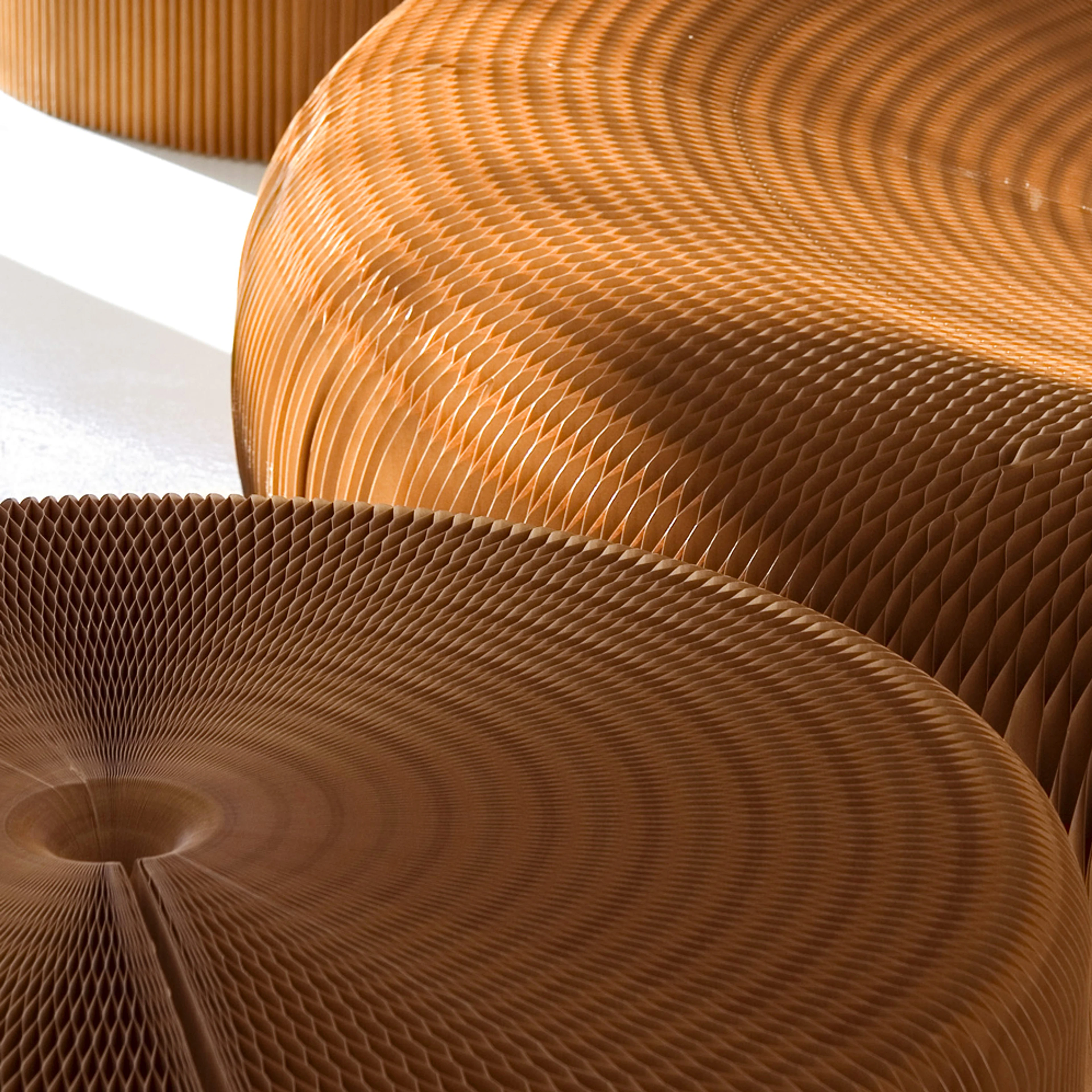 A close-up shot showcases the intricate honeycomb structure of several round, light brown, paper or cardboard fanning stools. The top surfaces reveal a mesmerizing radial pattern created by the folded material. Sunlight highlights the edges and textures of the stools, casting subtle shadows and emphasizing their depth and dimensionality. One stool in the foreground is a darker brown, while the ones behind it appear lighter, possibly due to lighting. A sliver of white floor is visible in the background.