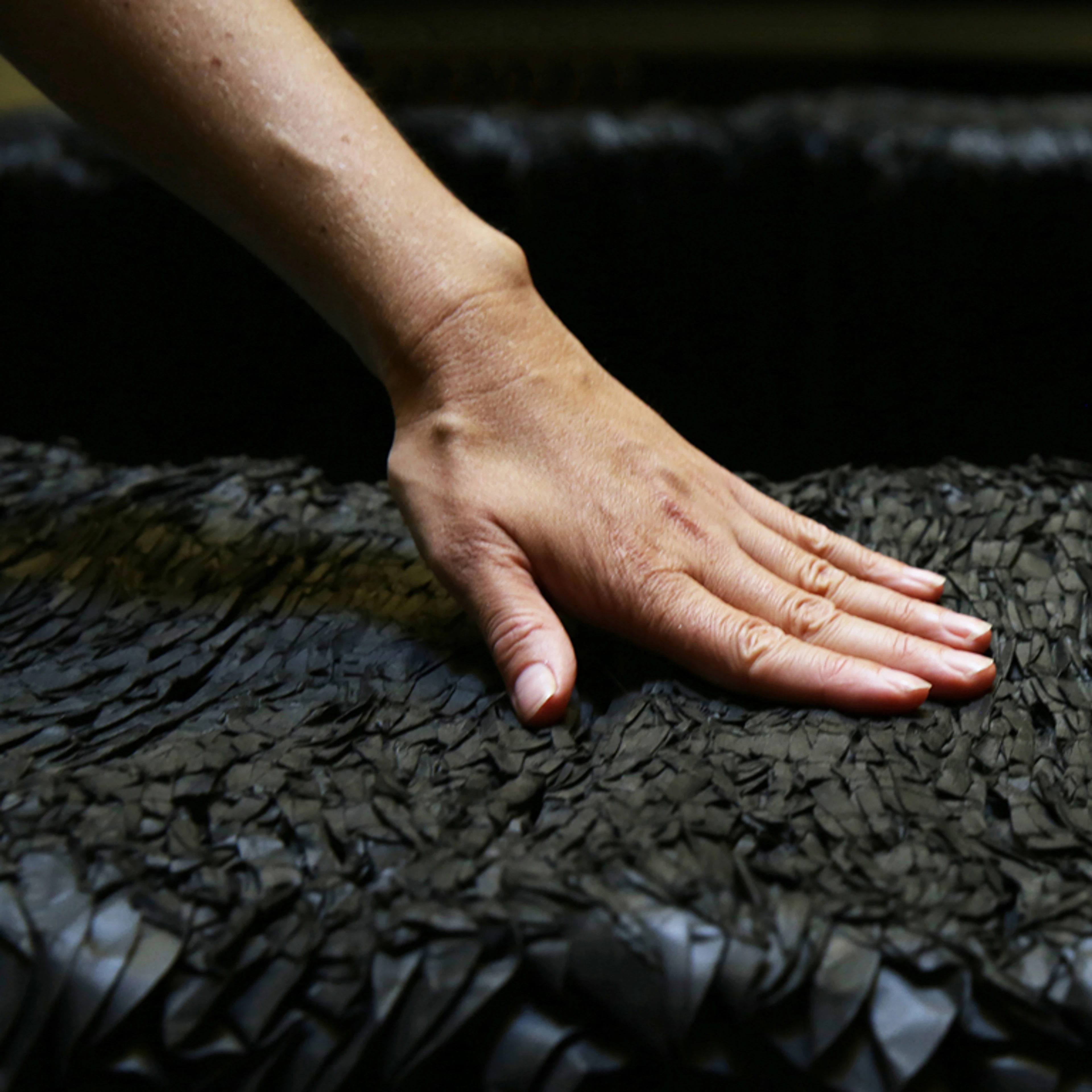 A close-up shot focuses on a person's left hand gently resting on the textured surface of a dark grey or black paper bench. The surface of the bench, originally likely smooth or uniformly patterned, now shows a compressed and variegated texture, with countless small, irregular, and somewhat flattened paper edges visible, indicating it has been sat upon and has conformed to use. The lighting highlights the details of the hand and the unique, worn texture of the bench against a dark, out-of-focus background.