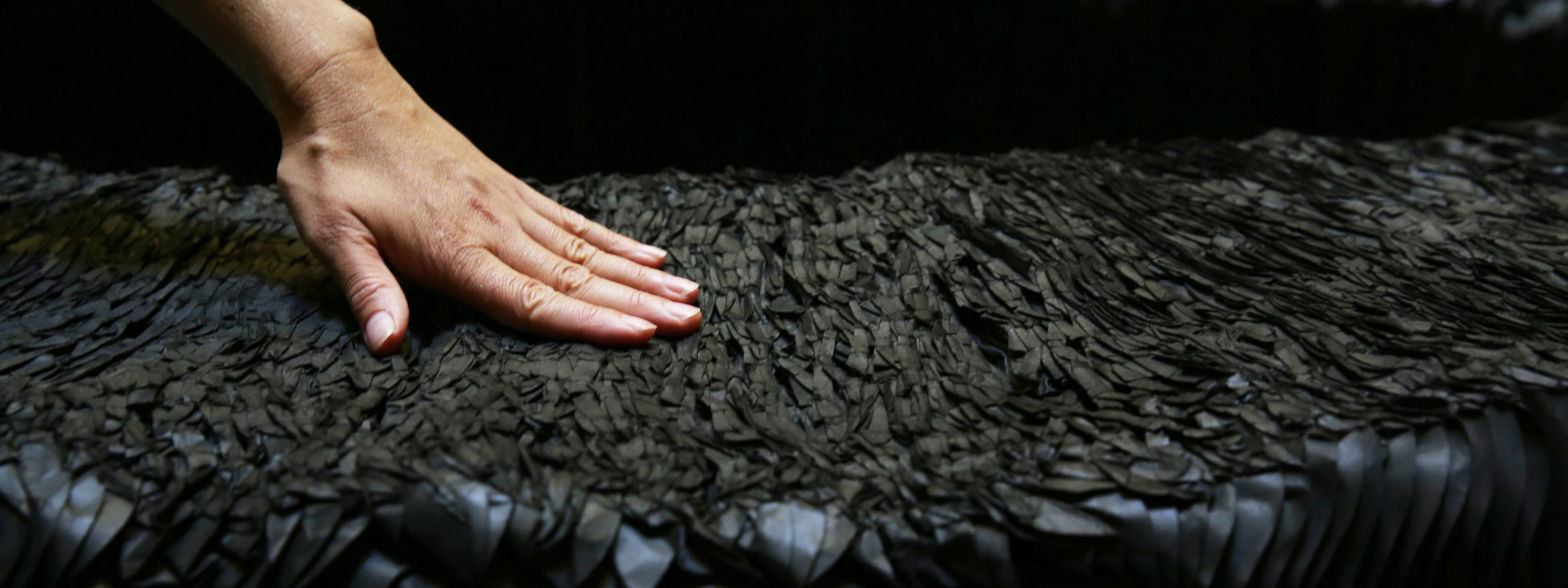A close-up shot focuses on a person's left hand gently resting on the textured surface of a dark grey or black paper bench. The surface of the bench, originally likely smooth or uniformly patterned, now shows a compressed and variegated texture, with countless small, irregular, and somewhat flattened paper edges visible, indicating it has been sat upon and has conformed to use. The lighting highlights the details of the hand and the unique, worn texture of the bench against a dark, out-of-focus background.
