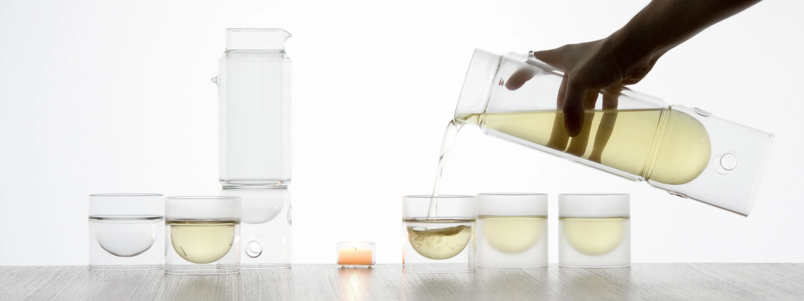 Against a brightly lit, white background, a hand is pouring a pale yellow liquid, likely tea, from a modern, clear glass carafe with a distinctive circular cutout handle into a small, clear, double-walled glass. The glass being filled is one of three identical glasses arranged in a row on a light wooden surface; the other two are already filled to different levels with the same pale yellow liquid. To the left, another similar clear glass contains clear water, and behind it stands a taller, cylindrical clear glass vessel, possibly a water filter or another carafe. A small, lit, peach-colored tea light candle sits on the wooden surface between the pouring action and the taller vessel. The image has a clean, minimalist aesthetic.