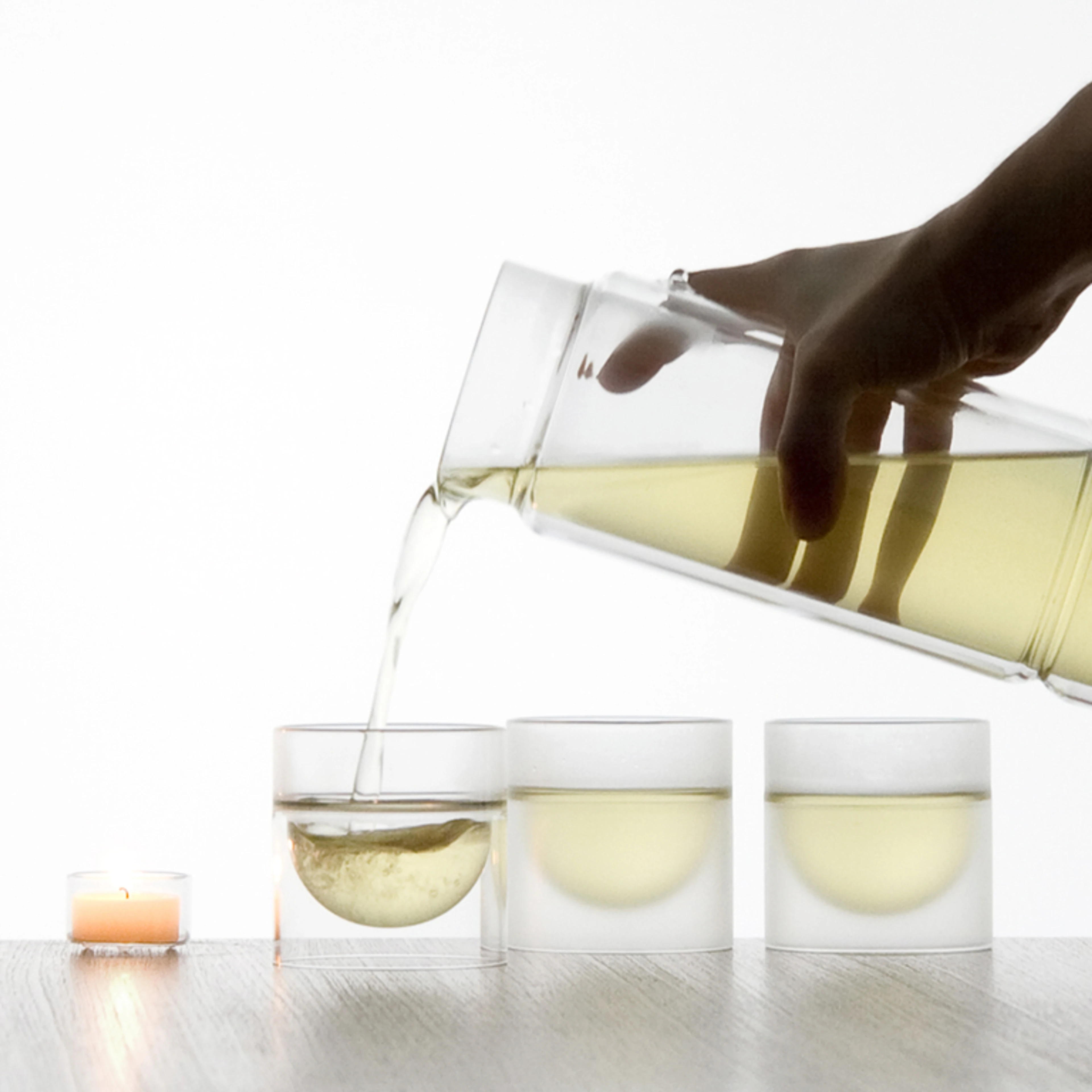 Against a brightly lit, white background, a hand is pouring a pale yellow liquid, likely tea, from a modern, clear glass carafe with a distinctive circular cutout handle into a small, clear, double-walled glass. The glass being filled is one of three identical glasses arranged in a row on a light wooden surface; the other two are already filled to different levels with the same pale yellow liquid. To the left, another similar clear glass contains clear water, and behind it stands a taller, cylindrical clear glass vessel, possibly a water filter or another carafe. A small, lit, peach-colored tea light candle sits on the wooden surface between the pouring action and the taller vessel. The image has a clean, minimalist aesthetic.