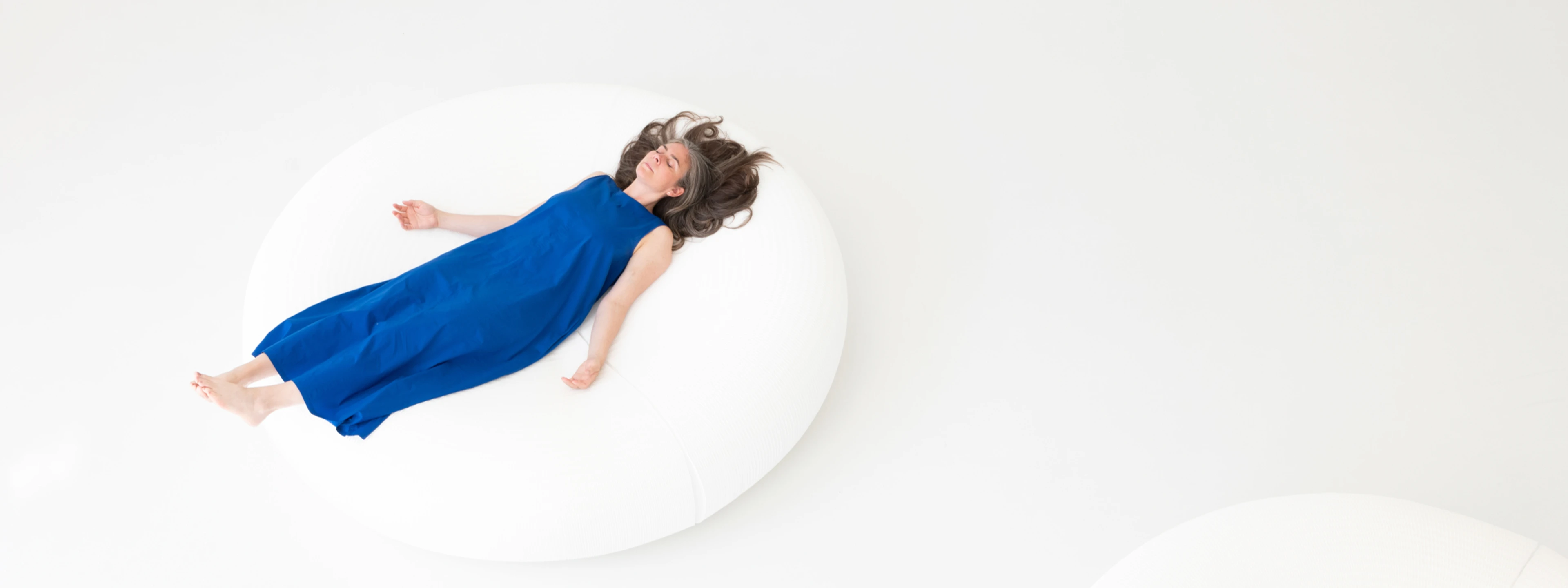 An overhead shot shows a person with long, dark hair, wearing a loose, sleeveless blue dress, lying on their back with arms slightly outstretched on a large, round, white cushion or beanbag. The background is entirely white, creating a minimalist and bright aesthetic.