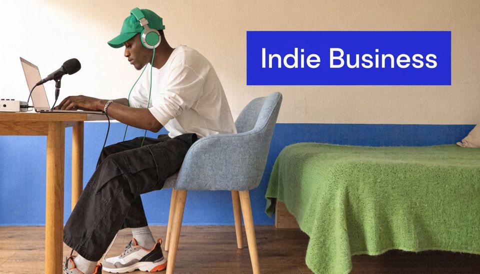 A young man wearing a green cap and headphones works on his laptop in a bedroom.