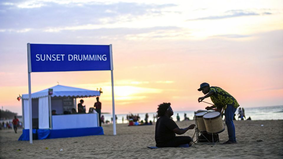 Two men playing drums on a sandy beach at sunset, beneath a "Sunset Drumming" sign.