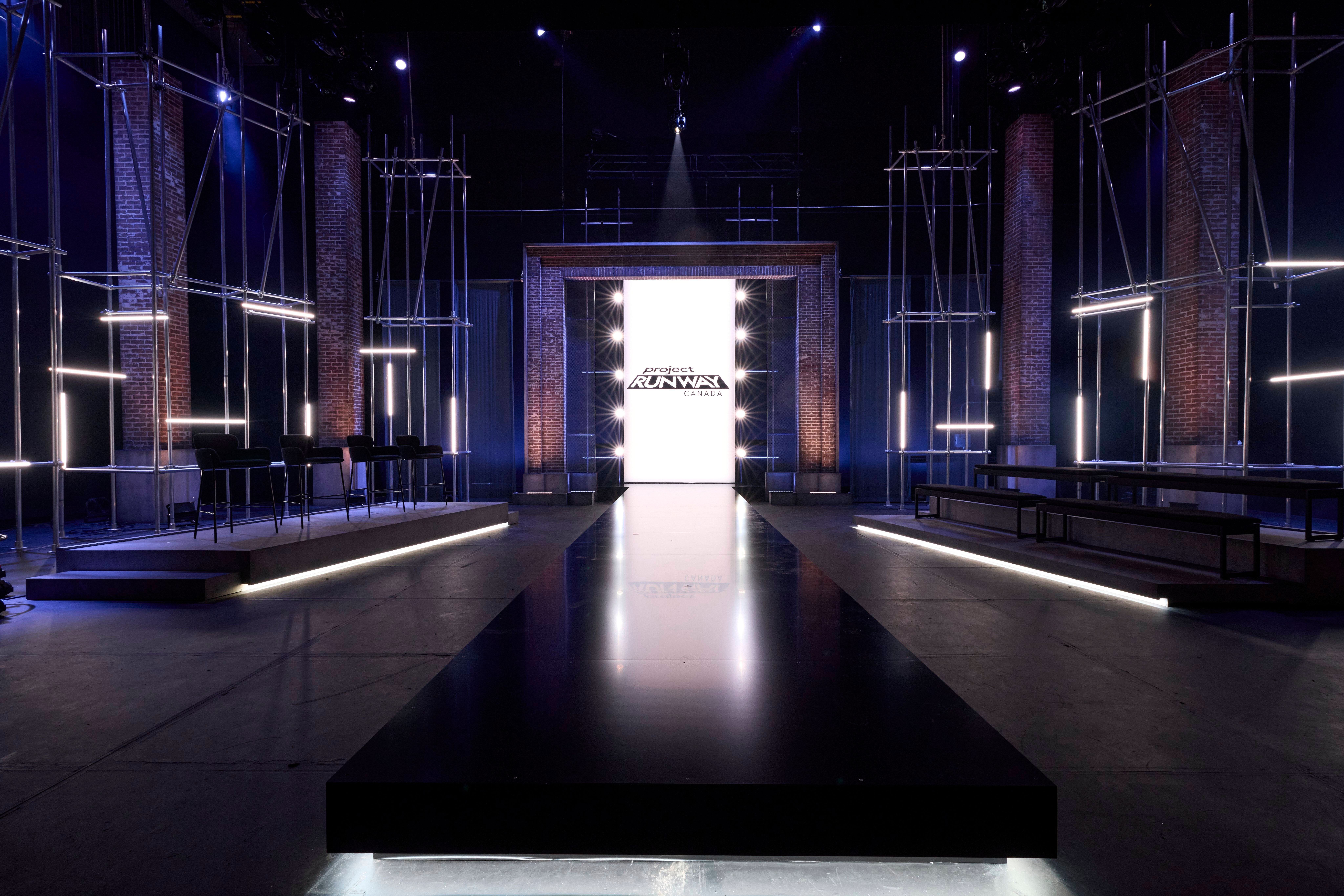 An empty, industrial-style fashion show set featuring a glossy black runway extending toward a brightly illuminated central doorway displaying the "Project Runway" logo, flanked by exposed brick pillars, metal scaffolding, and seating platforms.