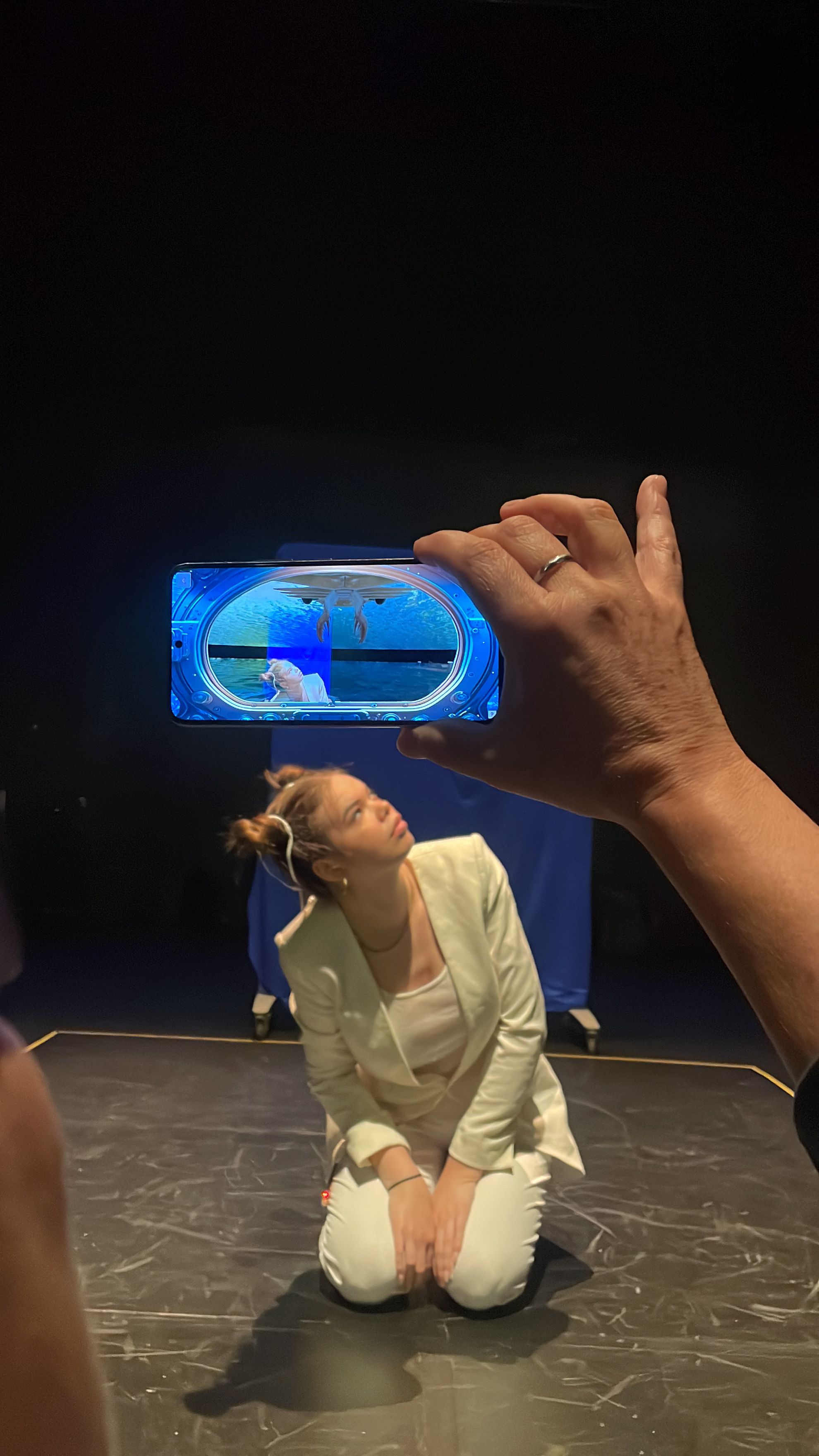 A person's hand holds a smartphone horizontally, framing a woman kneeling on a dark floor in a white blazer, while the phone screen displays an augmented reality overlay showing a virtual underwater scene with marine life.