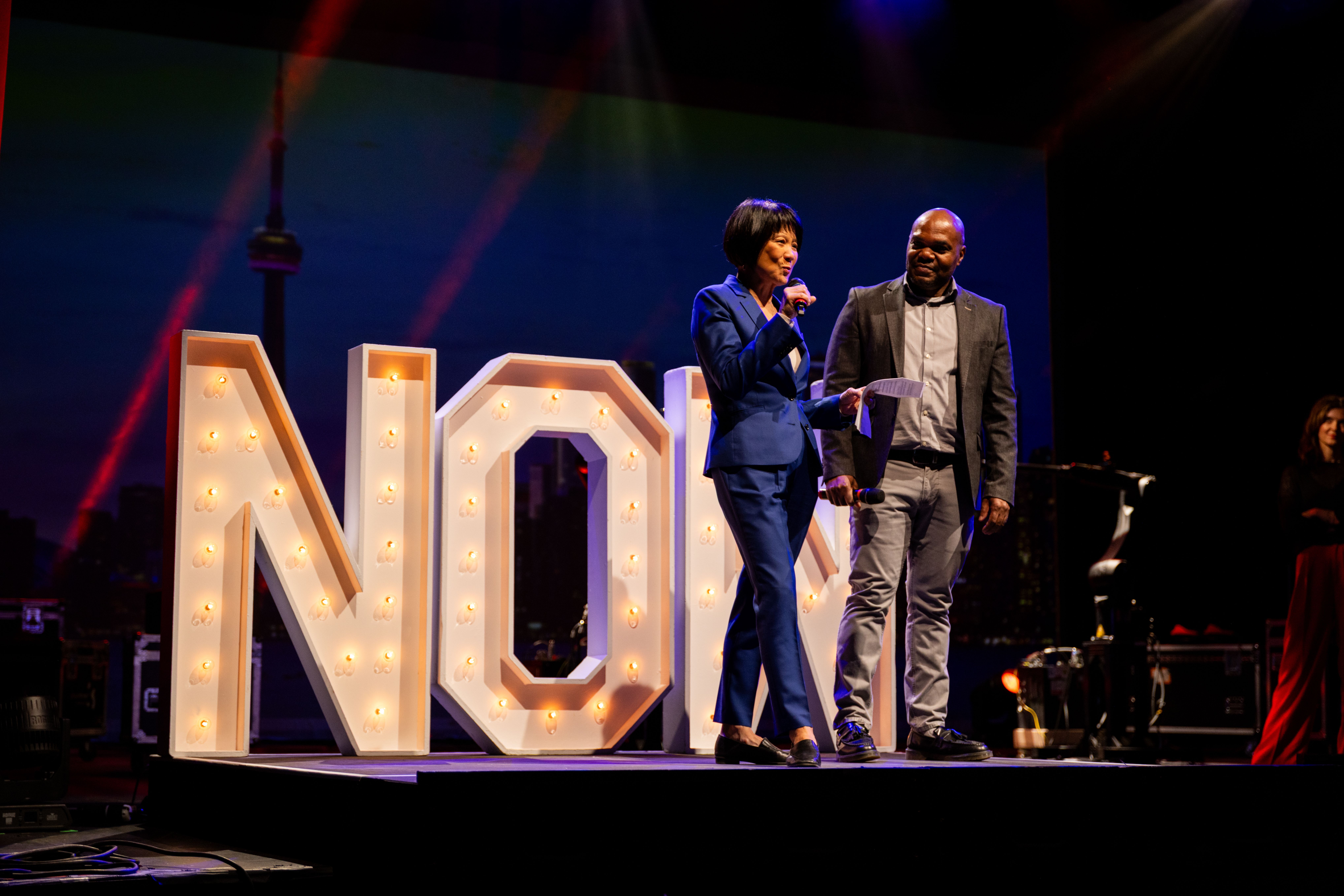 A woman and man speak onstage during an event, standing beside large illuminated letters in front of a cityscape backdrop.