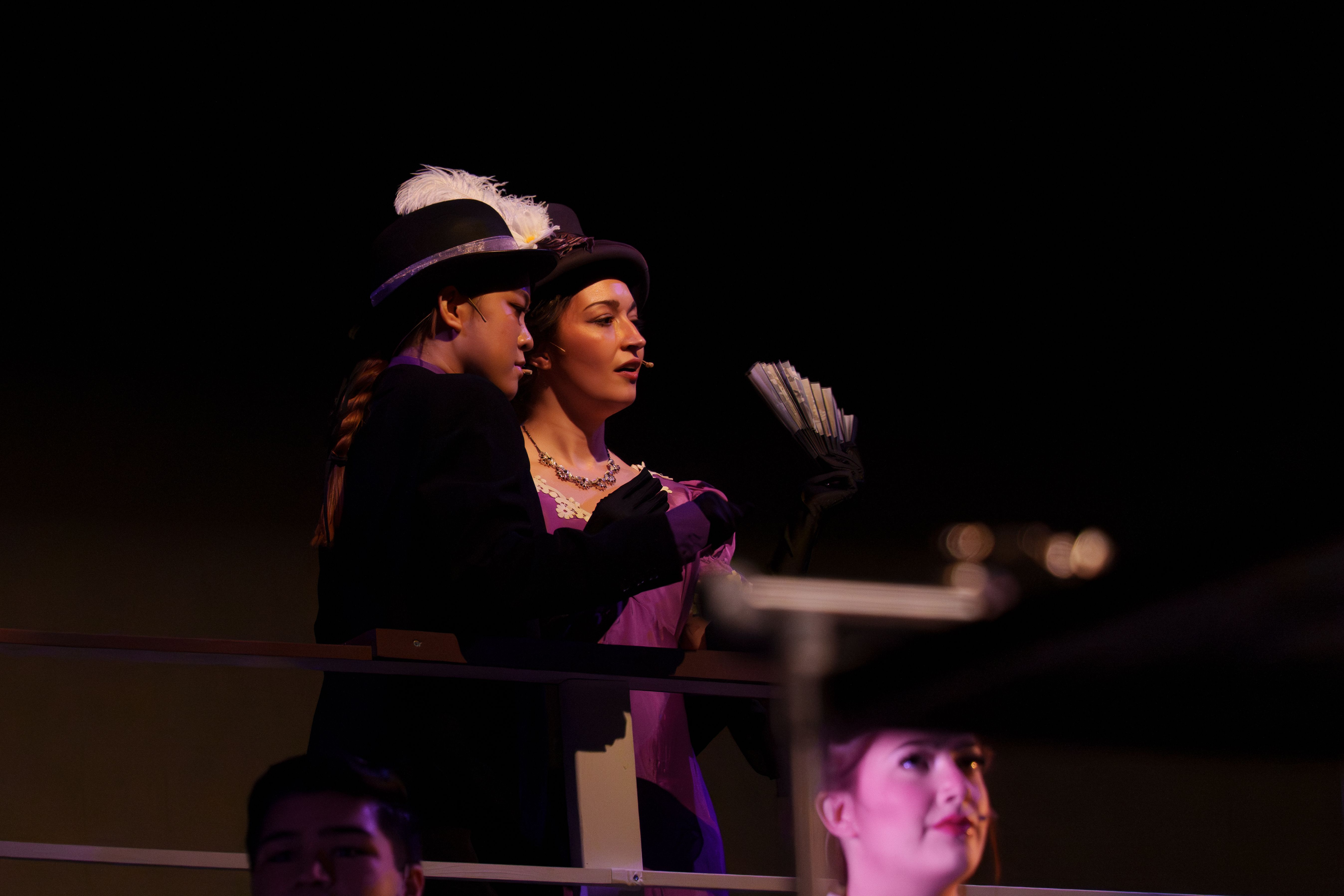 Two performers in theatrical costumes, including a woman wearing a white-feathered hat and a man in a purple vest, stand on a dark stage behind a railing, intently looking down toward an unseen area below while holding a fan.