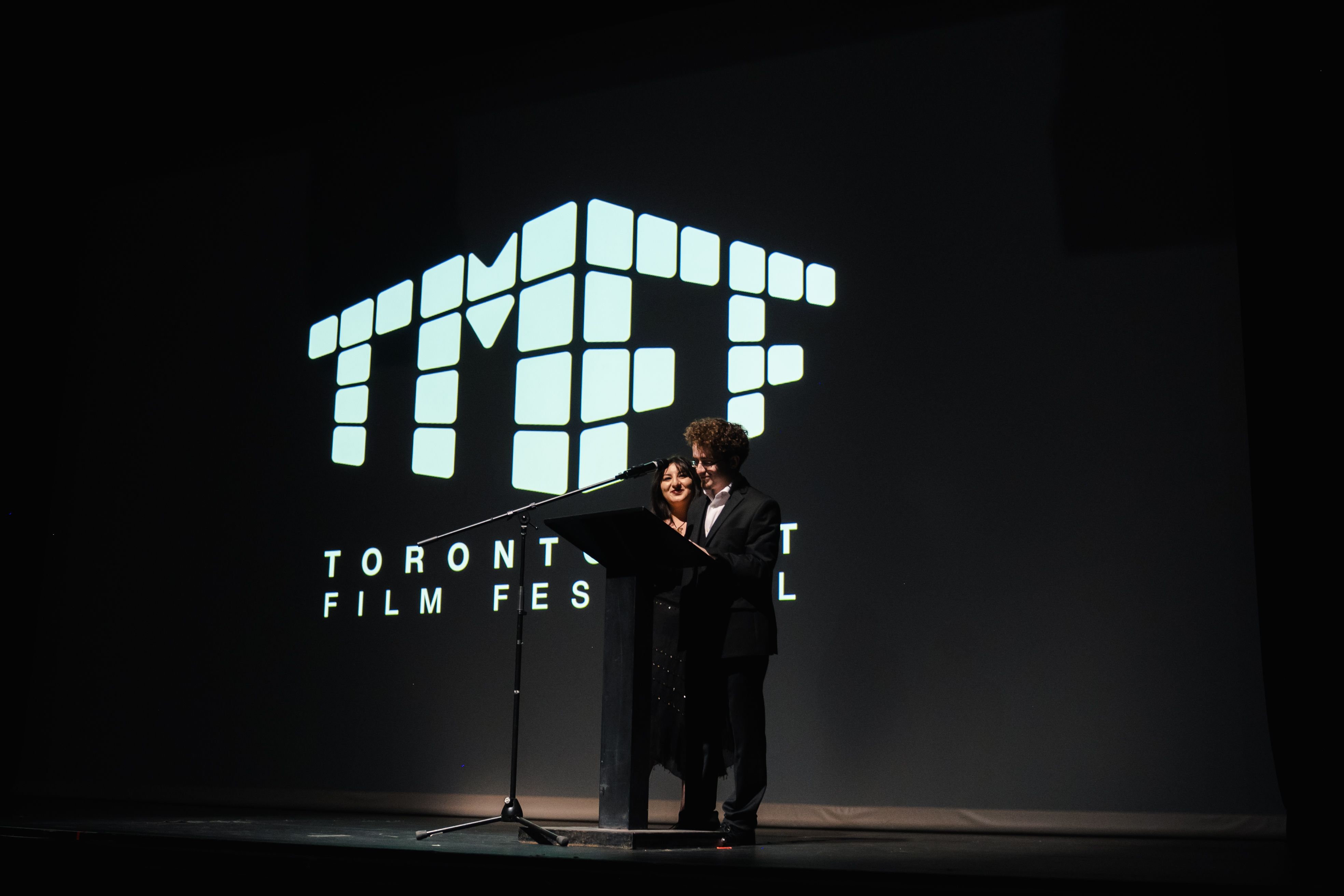 A man and a woman stand together smiling at a black podium on a dark stage, positioned in front of a large, illuminated screen displaying the "TIFF" cube logo and the text "TORONTO FILM FESTIVAL".