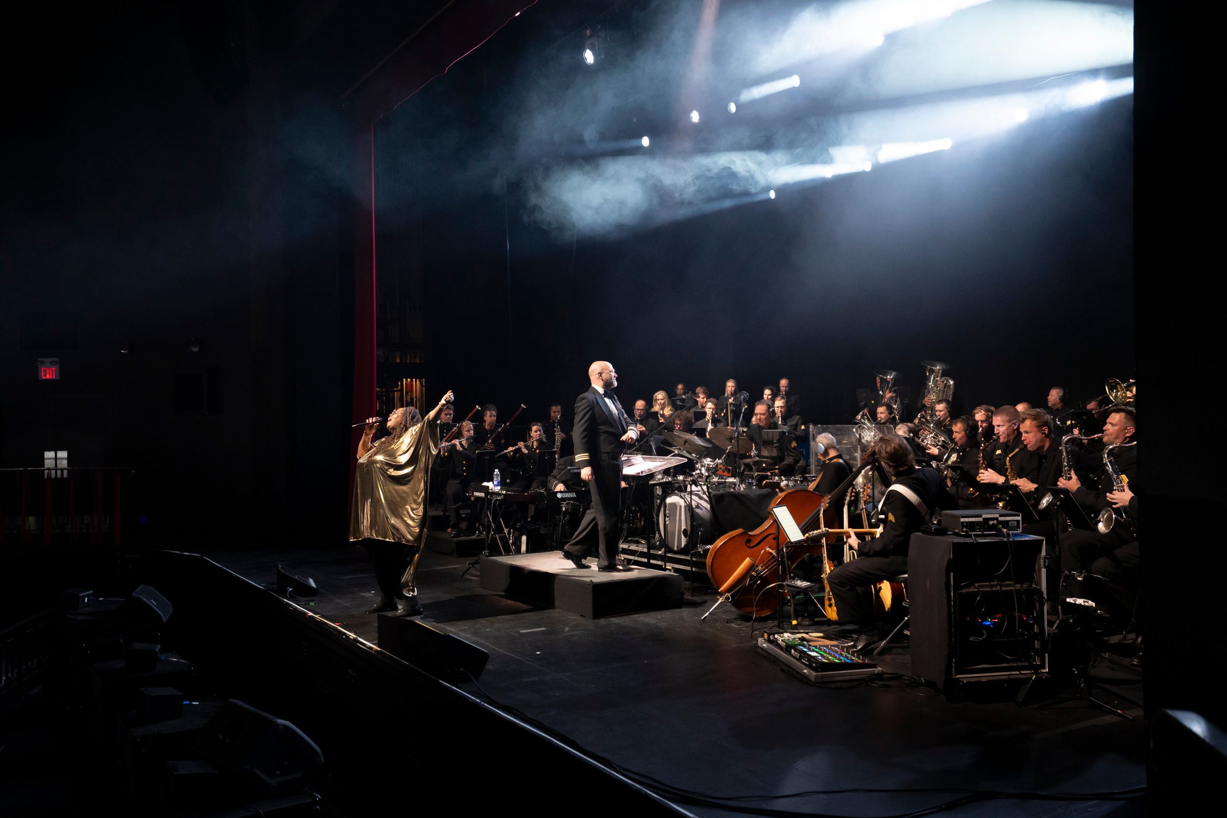 A theatrical concert performance taking place on a dark stage, where a singer in a flowing metallic gold dress stands with one arm raised, performing alongside a conductor in a dark uniform and a large seated orchestra, all illuminated by dramatic beams of light shining through stage haze.