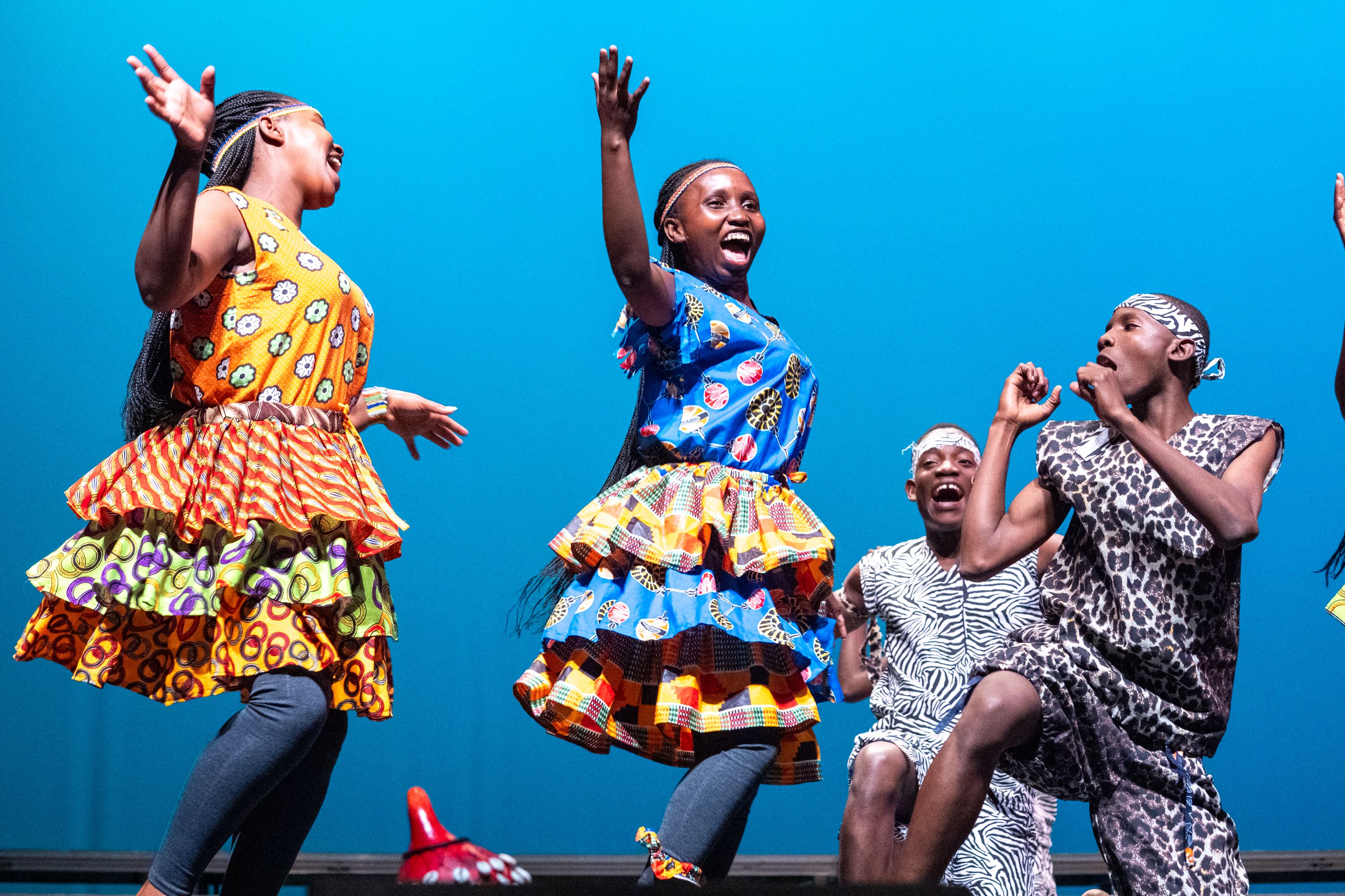 Four joyful performers in vibrant, patterned costumes—including layered dresses and animal-print outfits—are captured mid-dance against a bright blue backdrop, with three of them enthusiastically raising their hands and smiling.