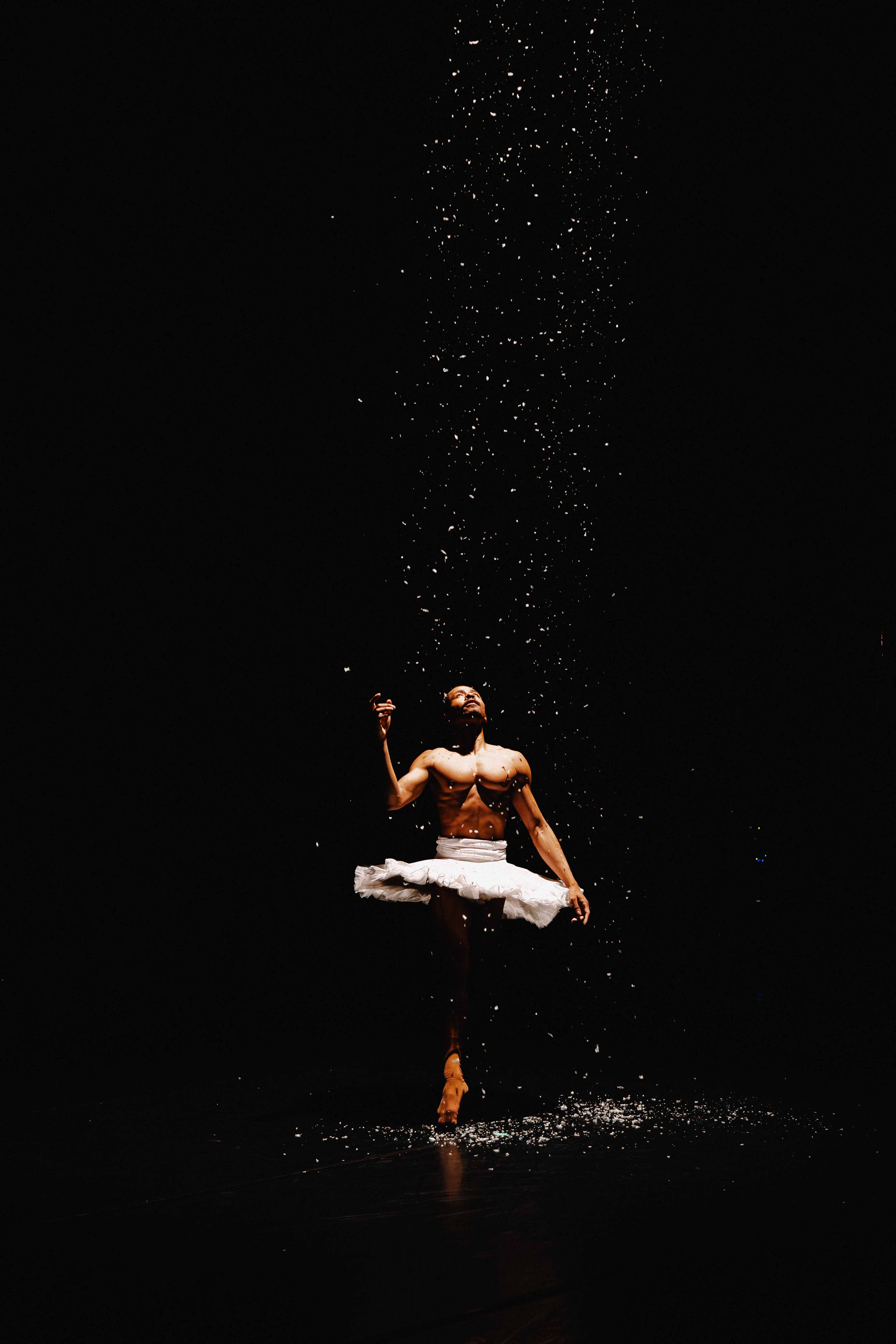 A shirtless male ballet dancer stands en pointe in a white tutu against a stark black backdrop, gazing upward as a shower of sparkling dust falls around him.