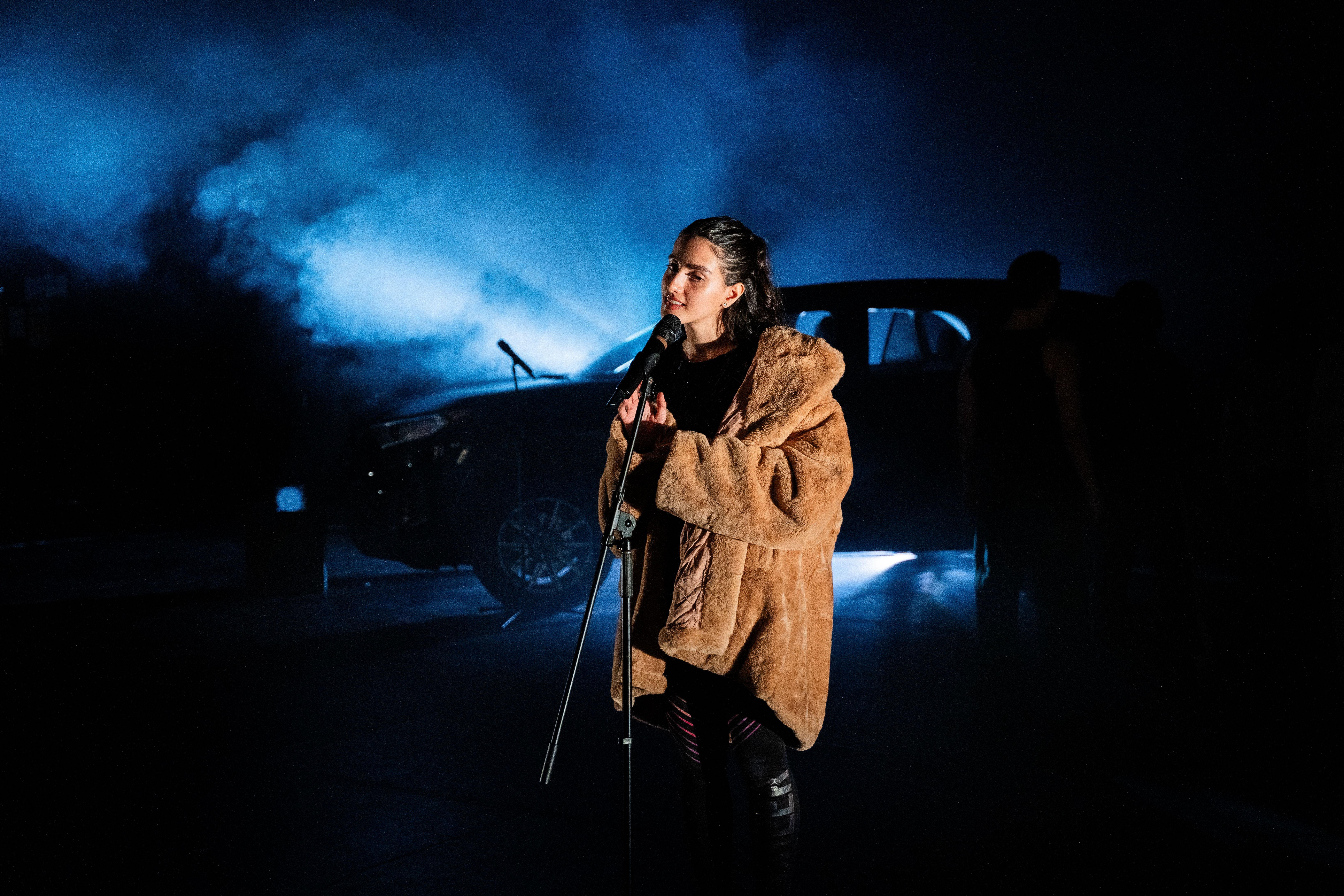A woman in a brown faux-fur coat sings into a microphone on a stand in a dark, smoky outdoor setting, illuminated by dramatic blue light with a parked car and a shadowy figure in the background.