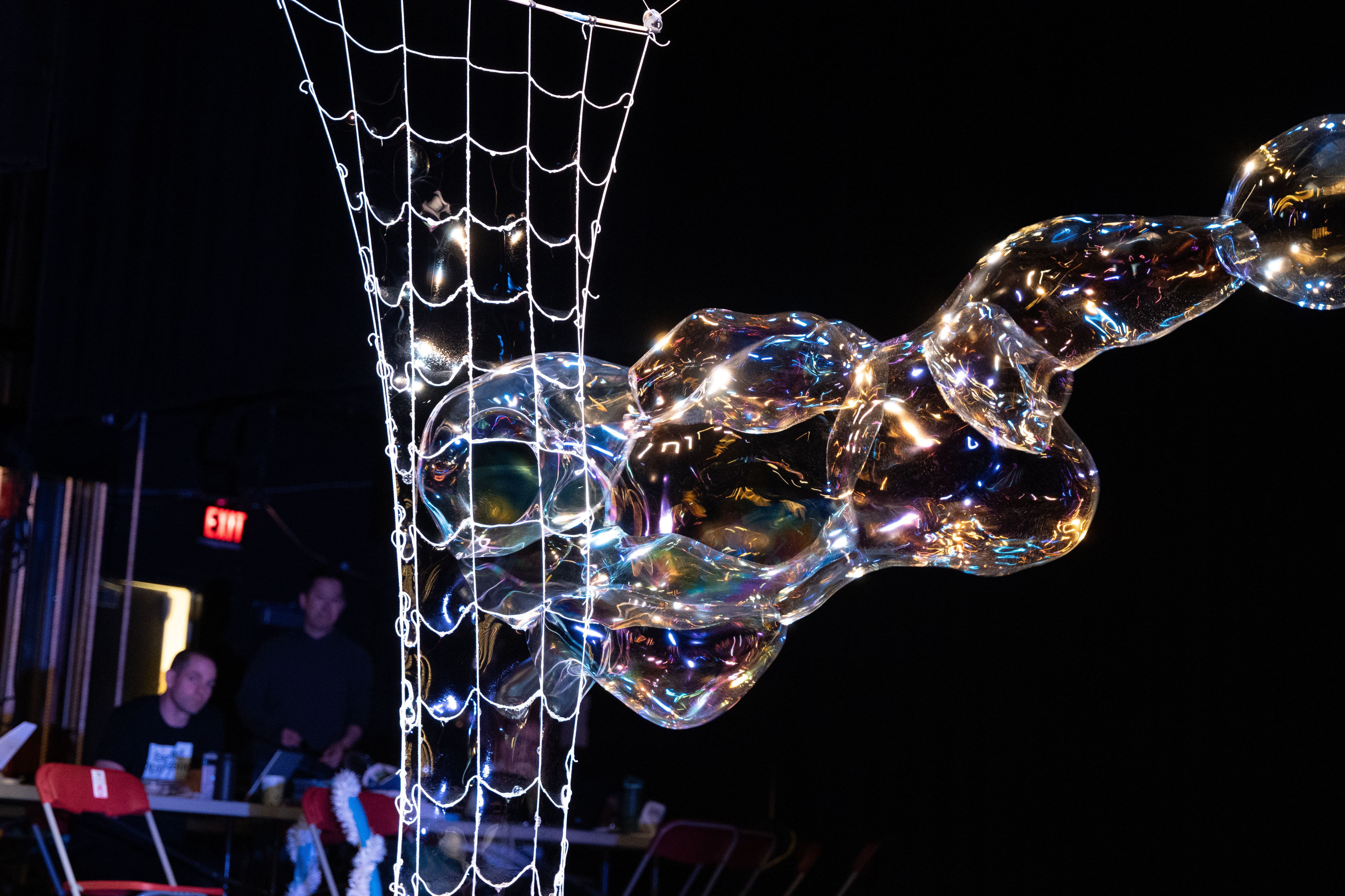 A large, iridescent bubble structure stretches outward from a white string net in a dark indoor space, with blurred figures and a glowing exit sign visible in the background.